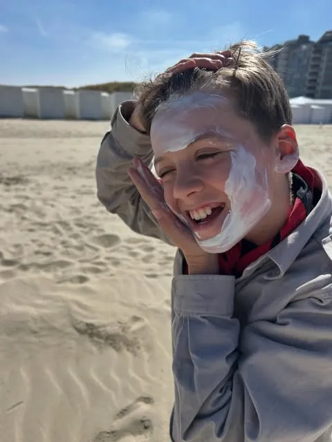 Een jongen met een dikke laag zonnecrème op zijn gezicht lacht op het strand, met strandcabines op de achtergrond.