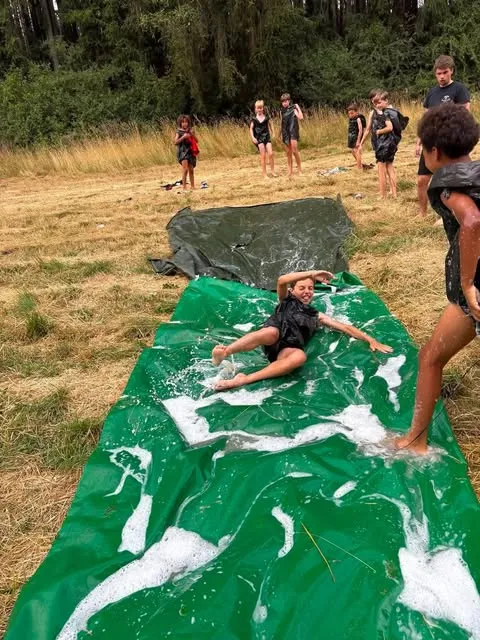 Een lid glijdt lachend over een nat, ingezeept zeil in het gras, terwijl anderen toekijken.